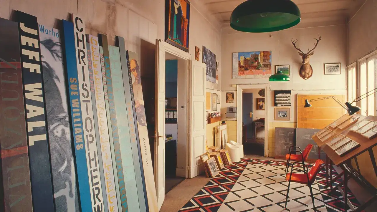 Estudio de dise&ntilde;o interiores carrera. Lienzos verticales con nombres de artistas y obras, un caballete de dibujo y alfombra geom&eacute;trica.