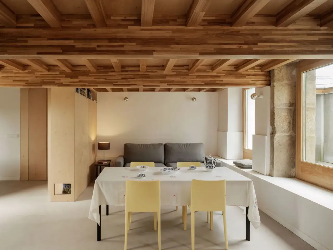 Un espacio acogedor con arquitectura y dise&ntilde;o de interiores modernos. Techo de madera, sof&aacute; gris, mesa con mantel y sillas amarillas.
