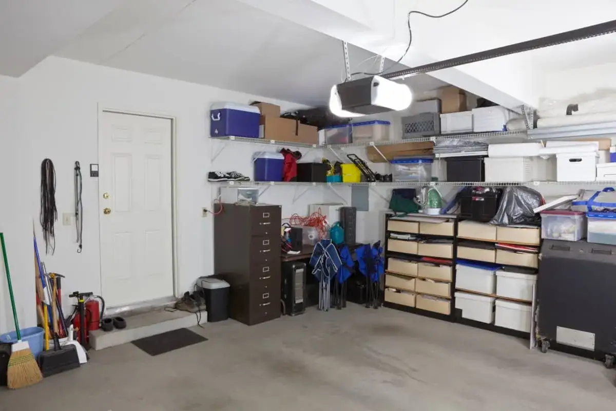 Estanter&iacute;as llenas de cajas y objetos, una puerta blanca y herramientas de limpieza. Un ejemplo de c&oacute;mo organizar un trastero para maximizar el espacio.