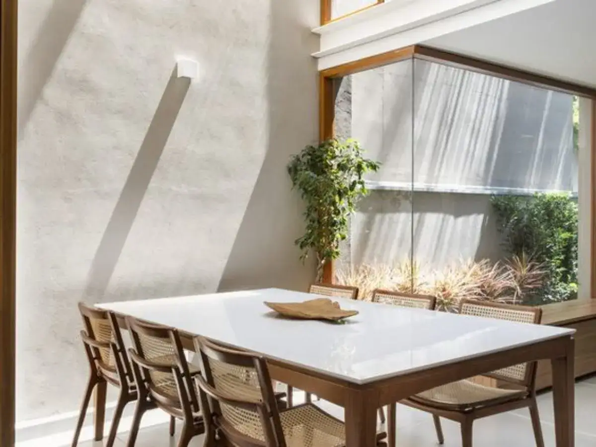 Comedor moderno con mesa blanca y sillas de mimbre, un ejemplo de casas de dise&ntilde;o interiores con luz natural y vegetaci&oacute;n exterior.