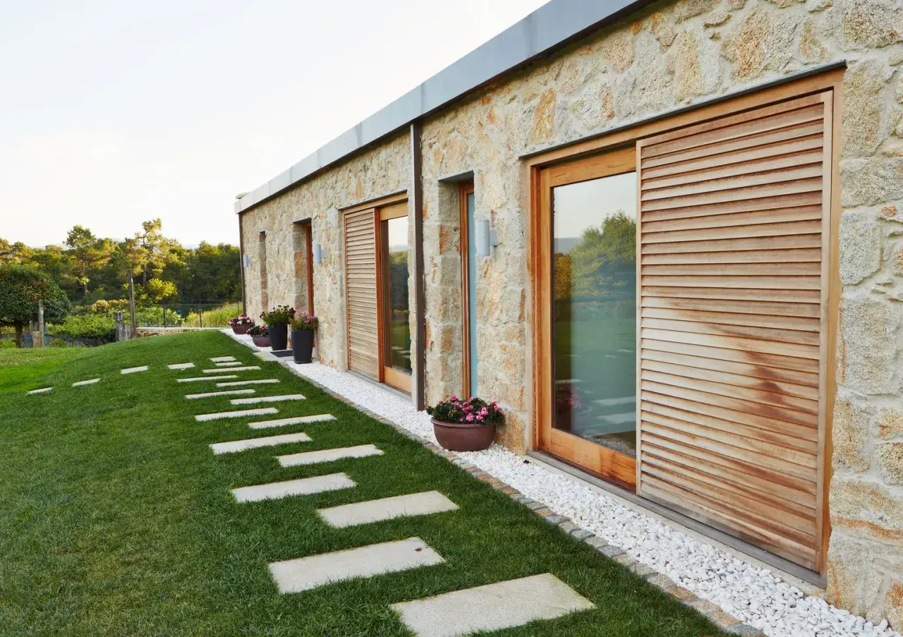 Acabados de paredes de piedra natural en una casa moderna, con puertas correderas de madera y un camino de losas en el c&eacute;sped.