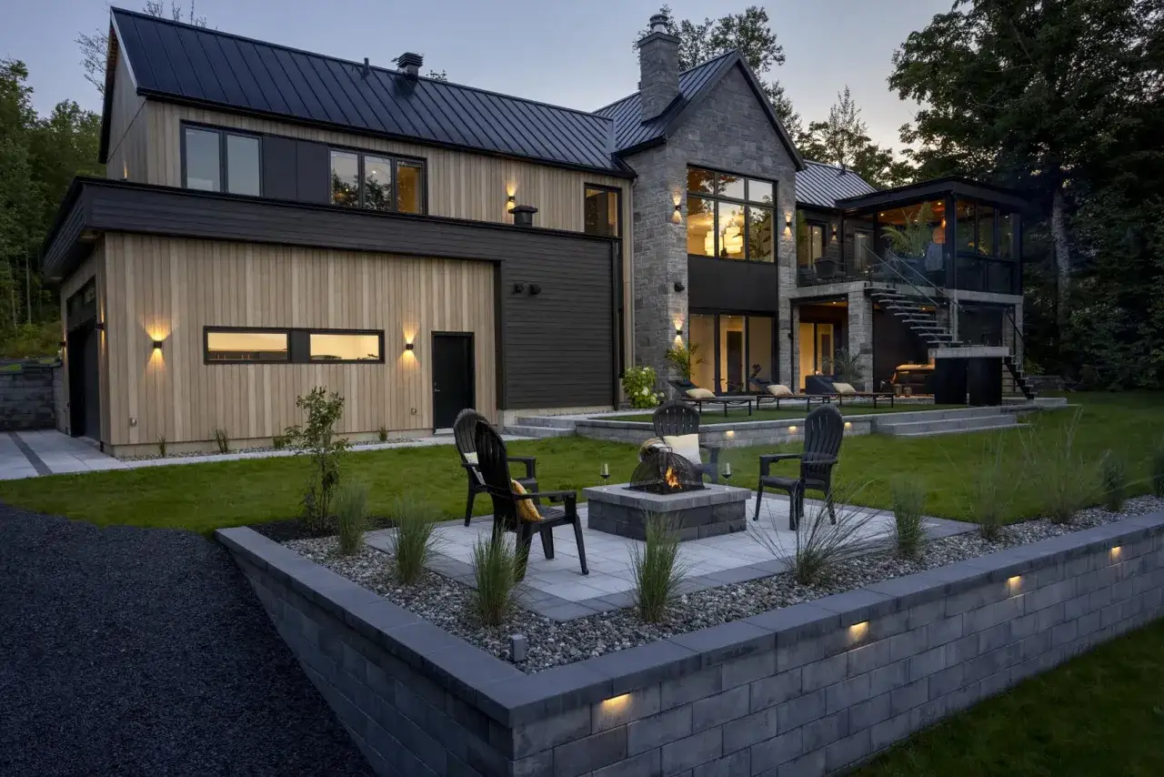 Modern house with stone and wood facade, featuring a fire pit area with chairs on tiered plots suelo and a lush green lawn.