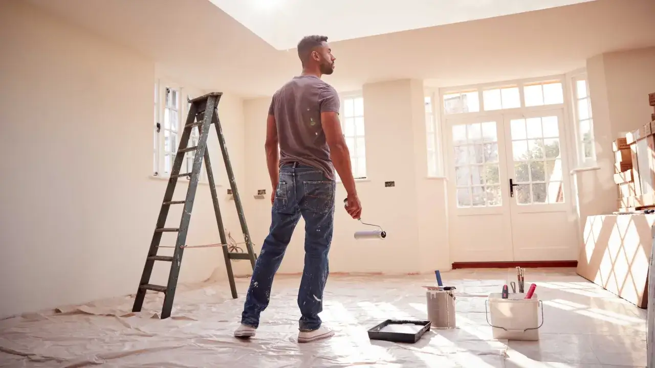 Hombre con rodillo listo para sanear pared en habitación con escalera y cubos de pintura.