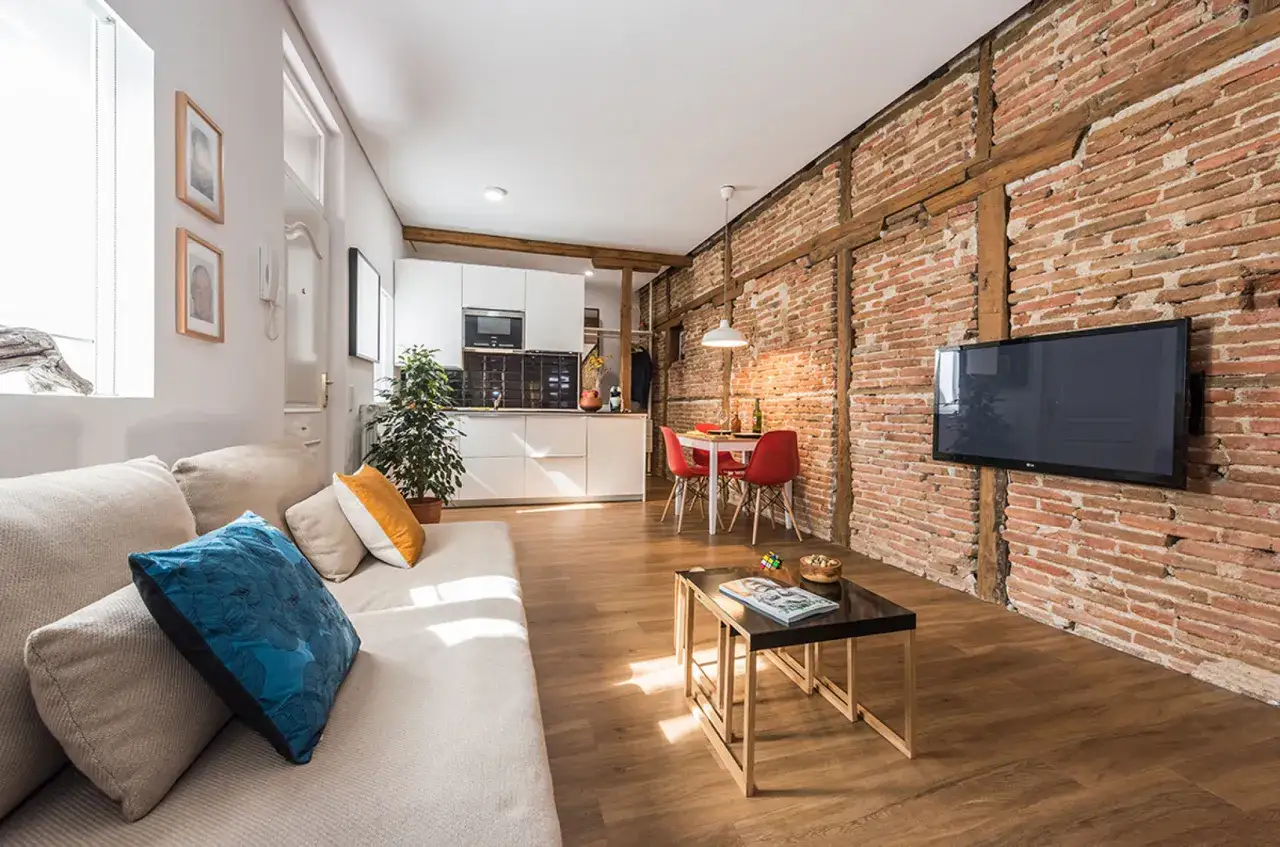 Sal&oacute;n moderno con sof&aacute; beige, mesa de centro, cocina blanca y una impresionante pared de ladrillo visto con vigas de madera.