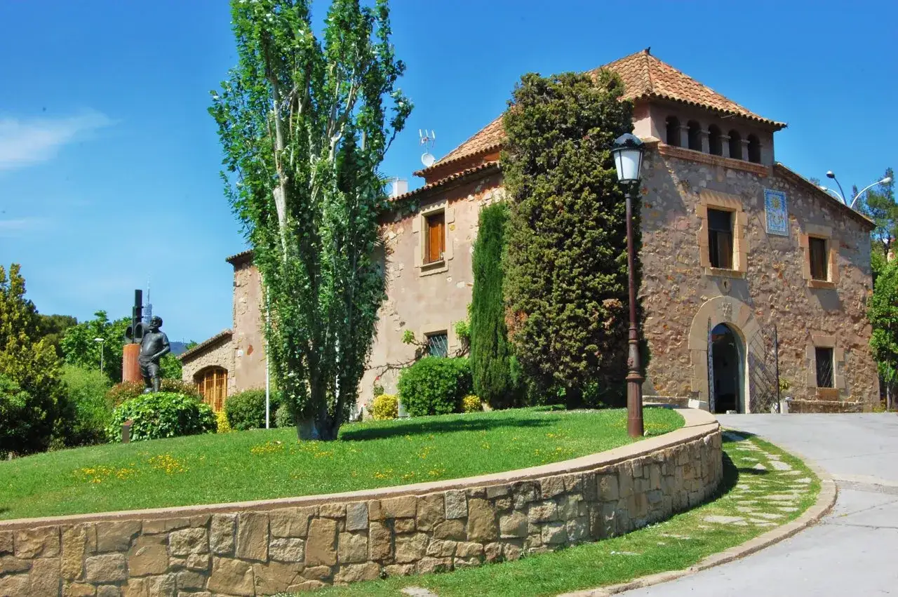 Edificio de arquitectura hist&oacute;rica con tejado de tejas, rodeado de vegetaci&oacute;n y un jard&iacute;n cuidado.