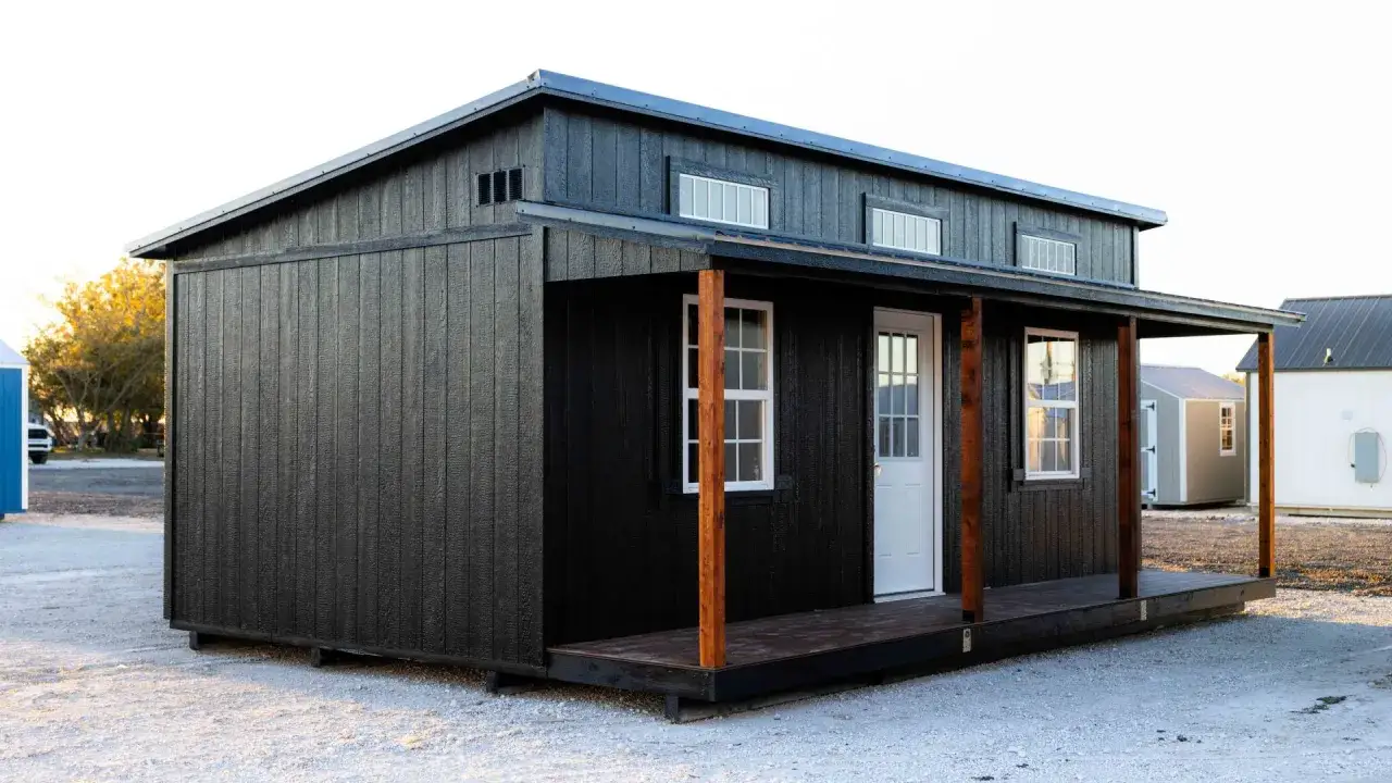 Casa prefabricada negra con porche y estructura de tejado a un agua. Ventanas rectangulares en el tejado y en la fachada.
