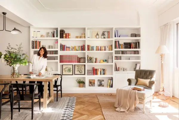 Una mujer sonríe junto a una mesa de comedor, rodeada de estanterías llenas de libros, un ejemplo de cómo los decoradores de interiores crean espacios acogedores.