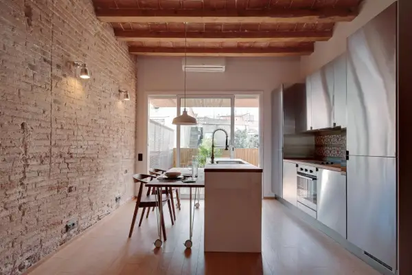 Cocina moderna con pared ladrillo visto, vigas de madera en el techo, isla central, electrodomésticos de acero inoxidable y mesa de comedor.