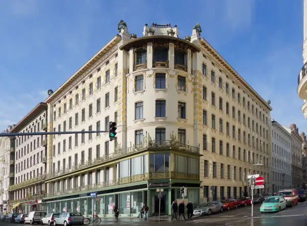 Edificio de esquina con detalles dorados, una obra de Otto Wagner. Fachada cream con ventanas oscuras y balcones verdes.