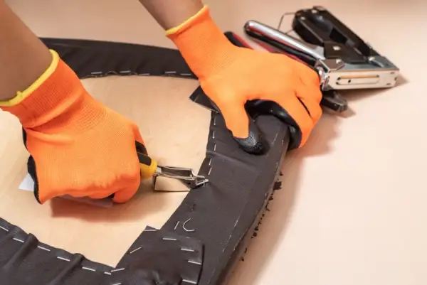 Manos con guantes naranjas usando una grapadora para tapizar una silla, fijando tela oscura a una base de madera.
