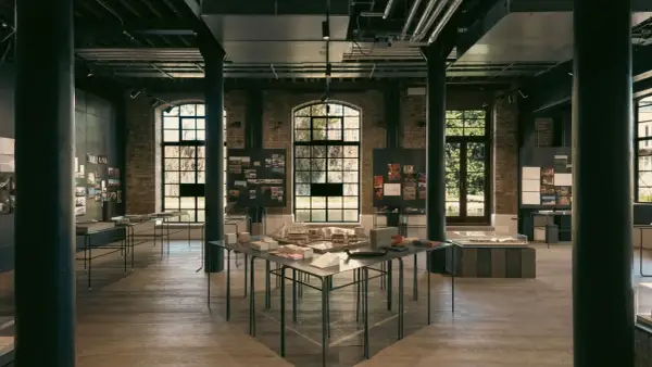 Exposición de arte y arquitectura en un espacio industrial con maquetas, planos y fotografías.