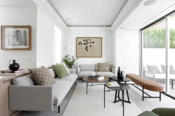 Elegante salón con sofás grises, mesas de centro negras y un banco de cuero marrón. El diseño de interiores y exteriores se fusiona con la luz natural que entra por los ventanales.