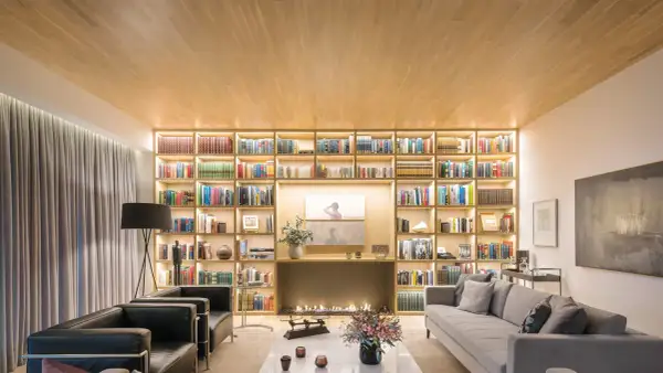 Un salón moderno con una gran librería iluminada, chimenea y sofás. Un diseñador de espacios ha creado un ambiente acogedor y elegante.
