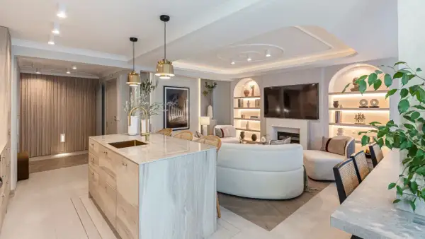 Cocina y sala de estar modernas con isla de cocina de madera, sofá curvo y estanterías iluminadas. La luz artificial crea un ambiente acogedor.